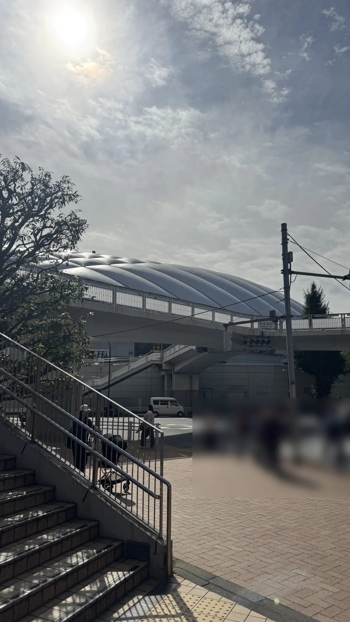 Tokyo Dome's white roof seen from the street outside Korakuen Station, with a stairway and a pedestrian bridge in the foreground