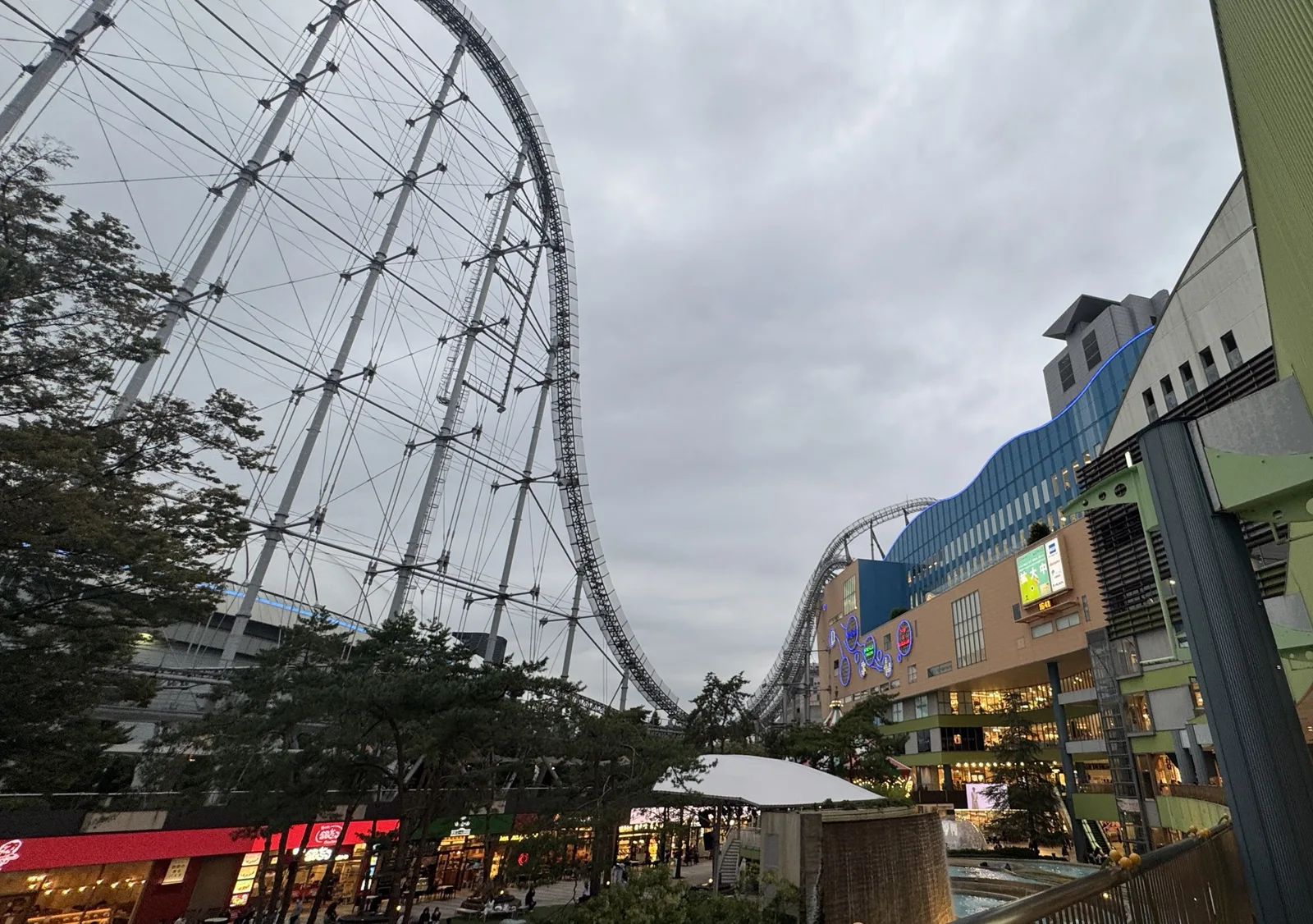 The LaQua building at dusk with lit shop fronts, a fountain in the courtyard and a roller coaster track running past the building