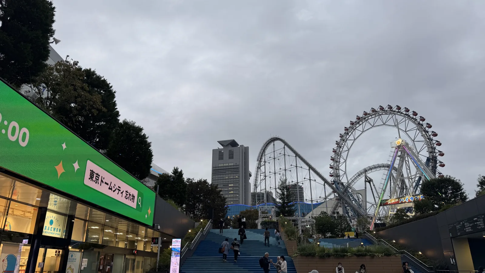 The wide blue staircase in Tokyo Dome City at dusk, with a ferris wheel, a roller coaster and the Tokyo Dome Hotel tower behind it